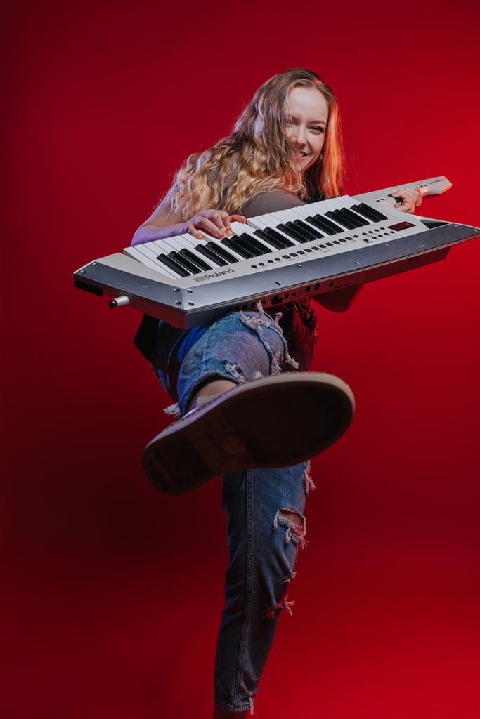 Woman with long blonde hair playing keytar with red background and kicking at camera.
