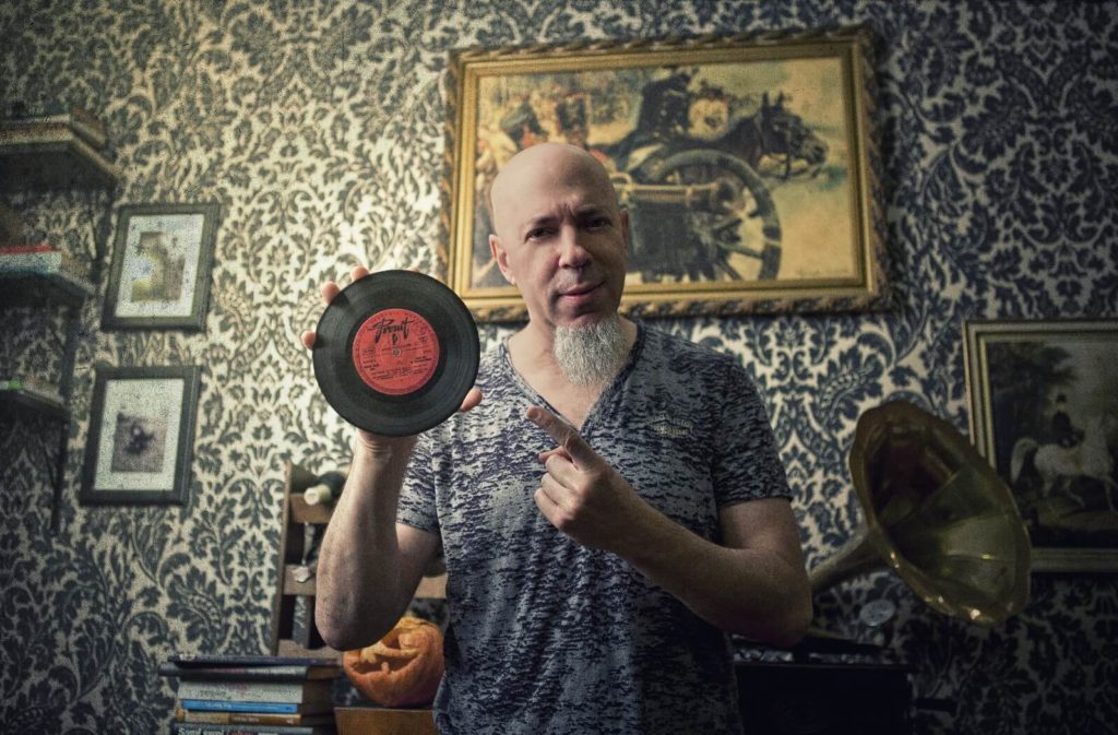 Man with goatee in grey tshirt holding up 7 inch record in room full of antiques.