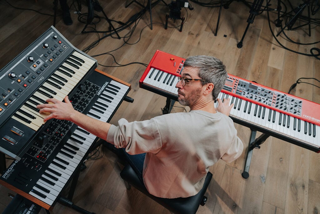 Shot from behind: Man with grey hair and glasses playing two stacked keyboards with left hand and one red keyboard (Nord Stage) with right hand.