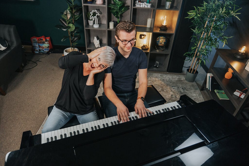 Sam and Lisa do piano improvisation exercises on the piano. Sam (brown hair, glasses) sits at piano playing on the right. Lisa (platinum short hair) holds her face tilted sideways in a 'wow' expression.