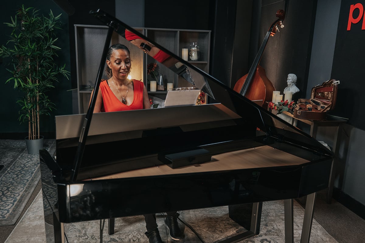 Victoria Theodore (woman in red dress) behind digital grand piano with open lid in studio with double bass and violin case.